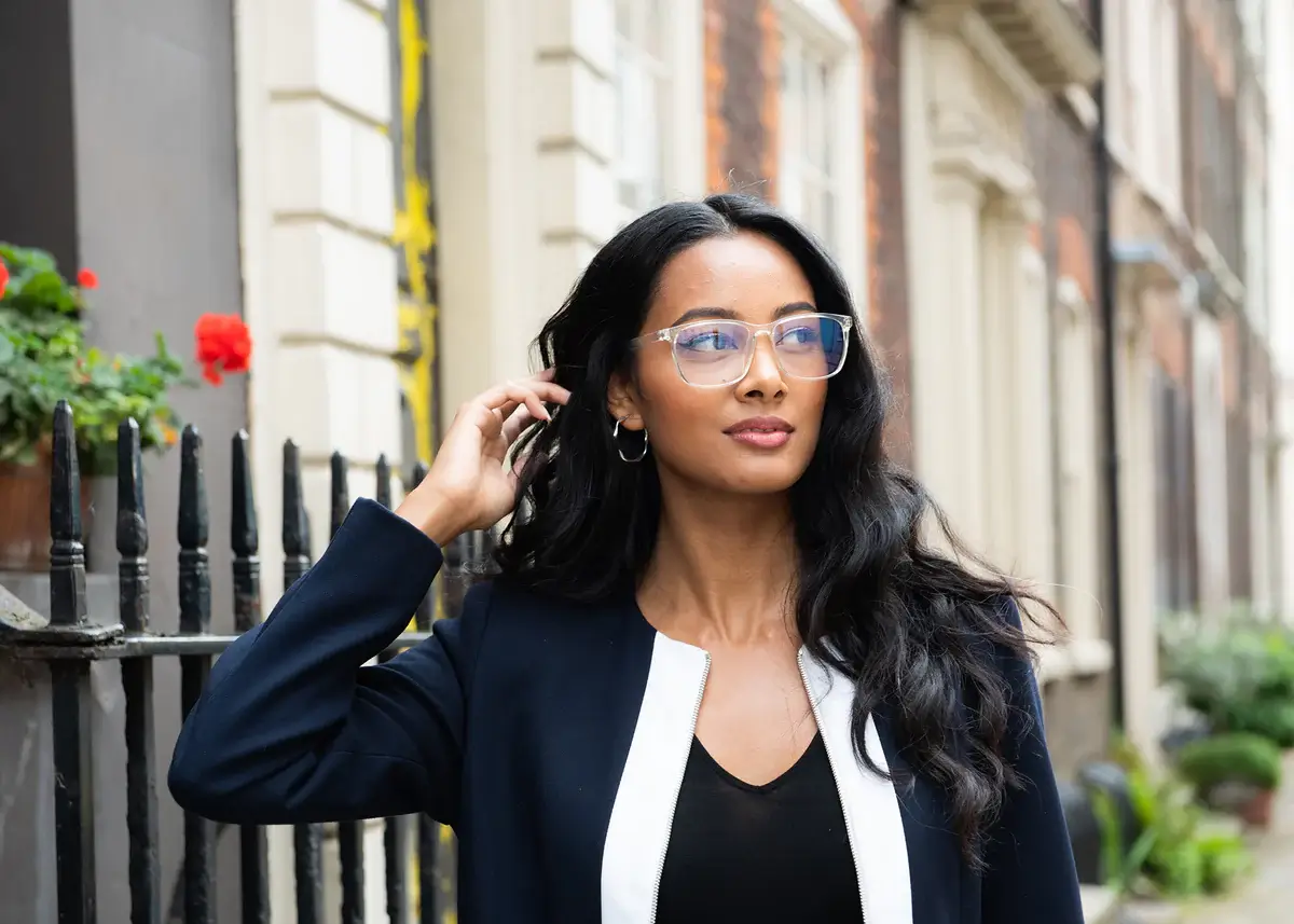 4_anti-blue-light-glasses_1200x Woman with long dark hair and clear-framed glasses stands on a city sidewalk beside a black iron fence, posing for the photo.