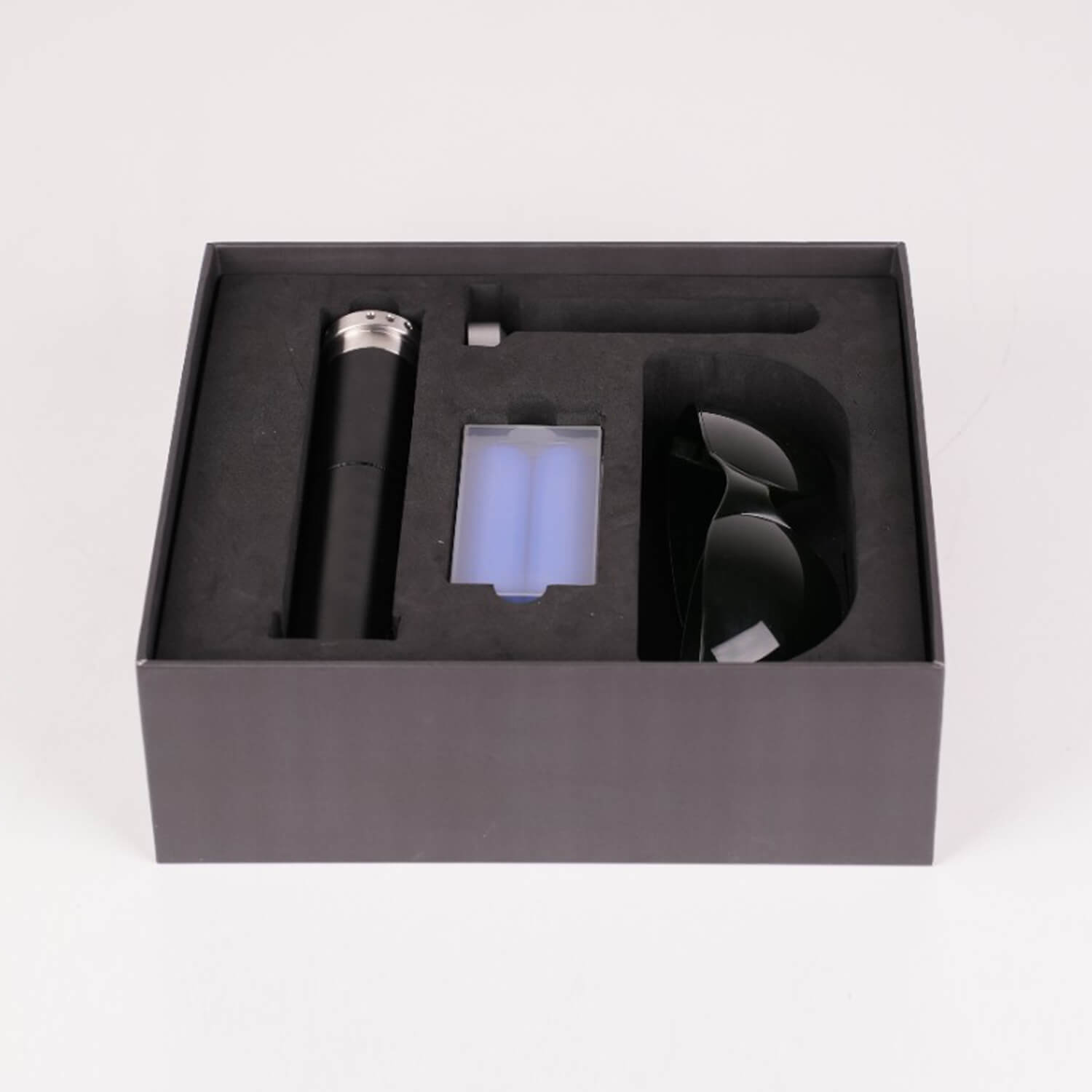 Open black foam insert box showing a cylindrical black device on the left, a blue battery pack in the center, and a curved black accessory on the right.