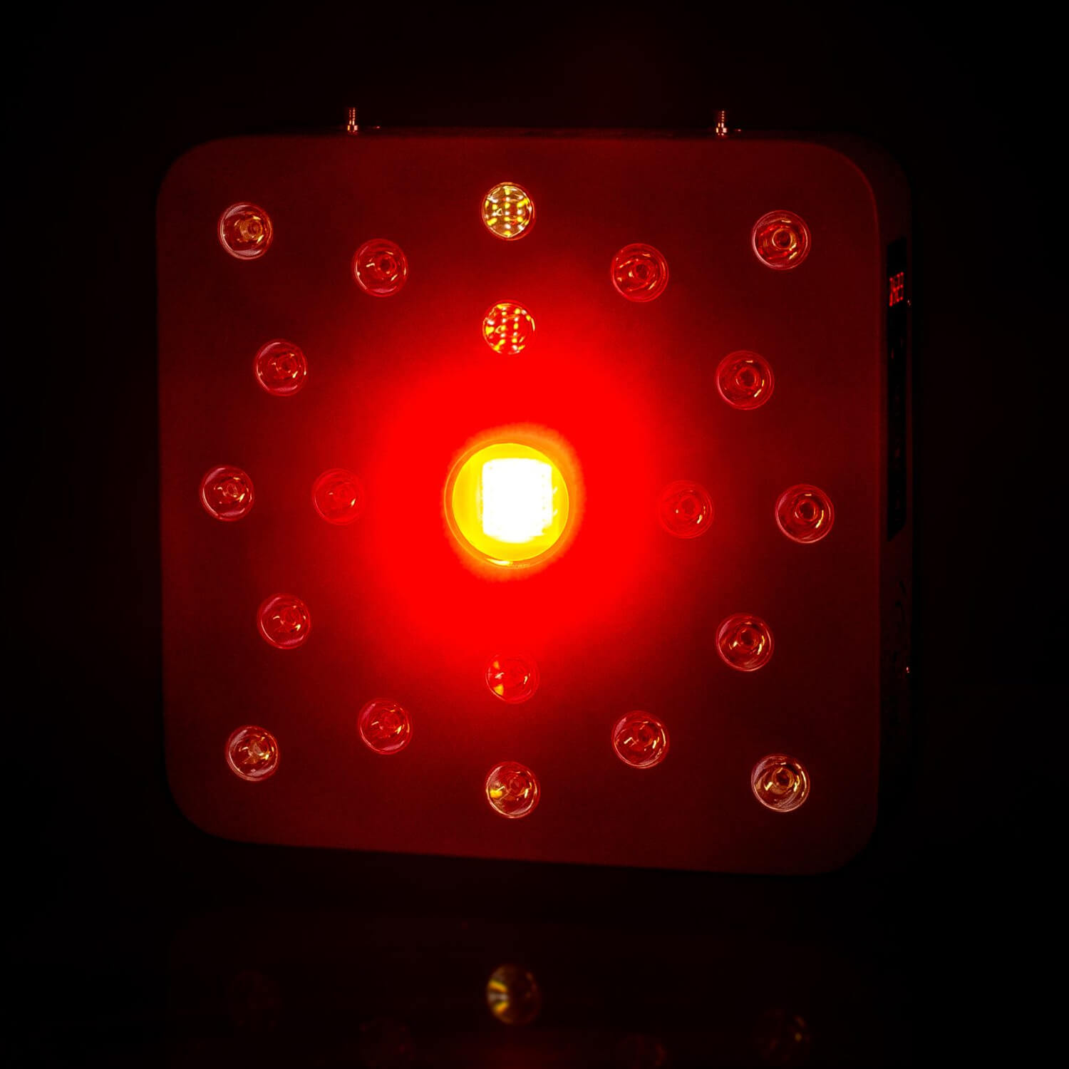 Red LED panel with multiple circular diodes surrounding a bright central LED, glowing red in darkness (grows light/therapy device).