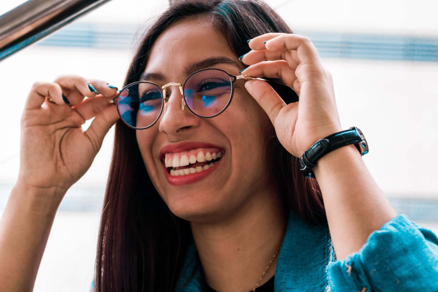 glasses-unsplash-scaled-1 Woman smiling while adjusting round glasses outdoors, wearing a teal jacket and a black watch on her left wrist.