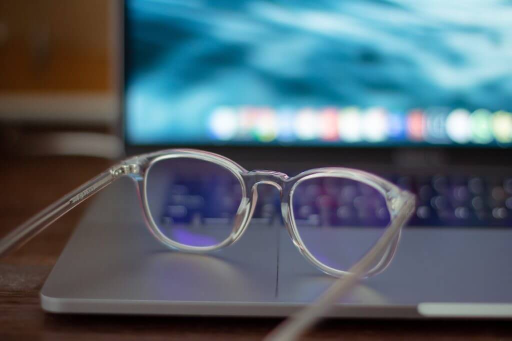 k8-r87zX1RWECQ-unsplash-1024×683-1 Clear glasses resting on a silver laptop with a blue blurred screen in the background.