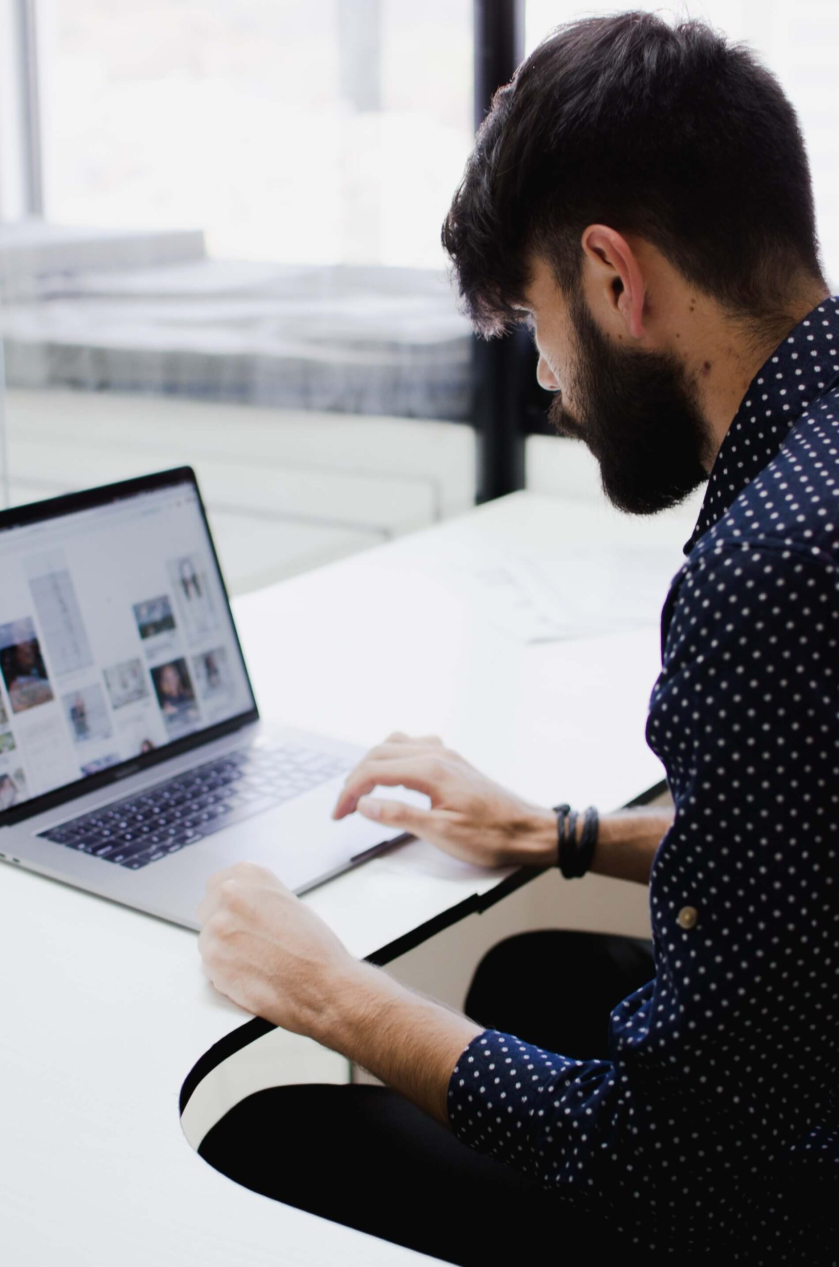miguelangel-miquelena-EJ3aJ9AuYtg-unsplash Bearded man in a navy polka-dot shirt typing on a laptop at a bright white desk near a window.