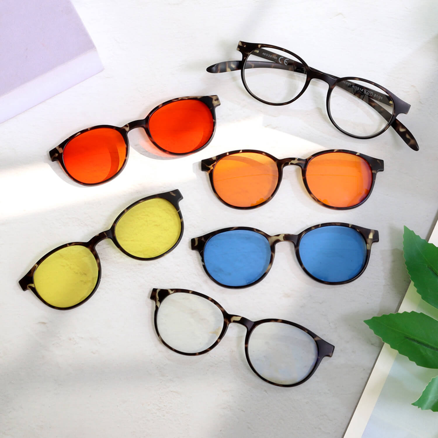 Six colorful sunglasses with tortoiseshell frames and orange, yellow, and blue lenses scattered on a white surface with a purple corner of a notebook and green leaves nearby.
