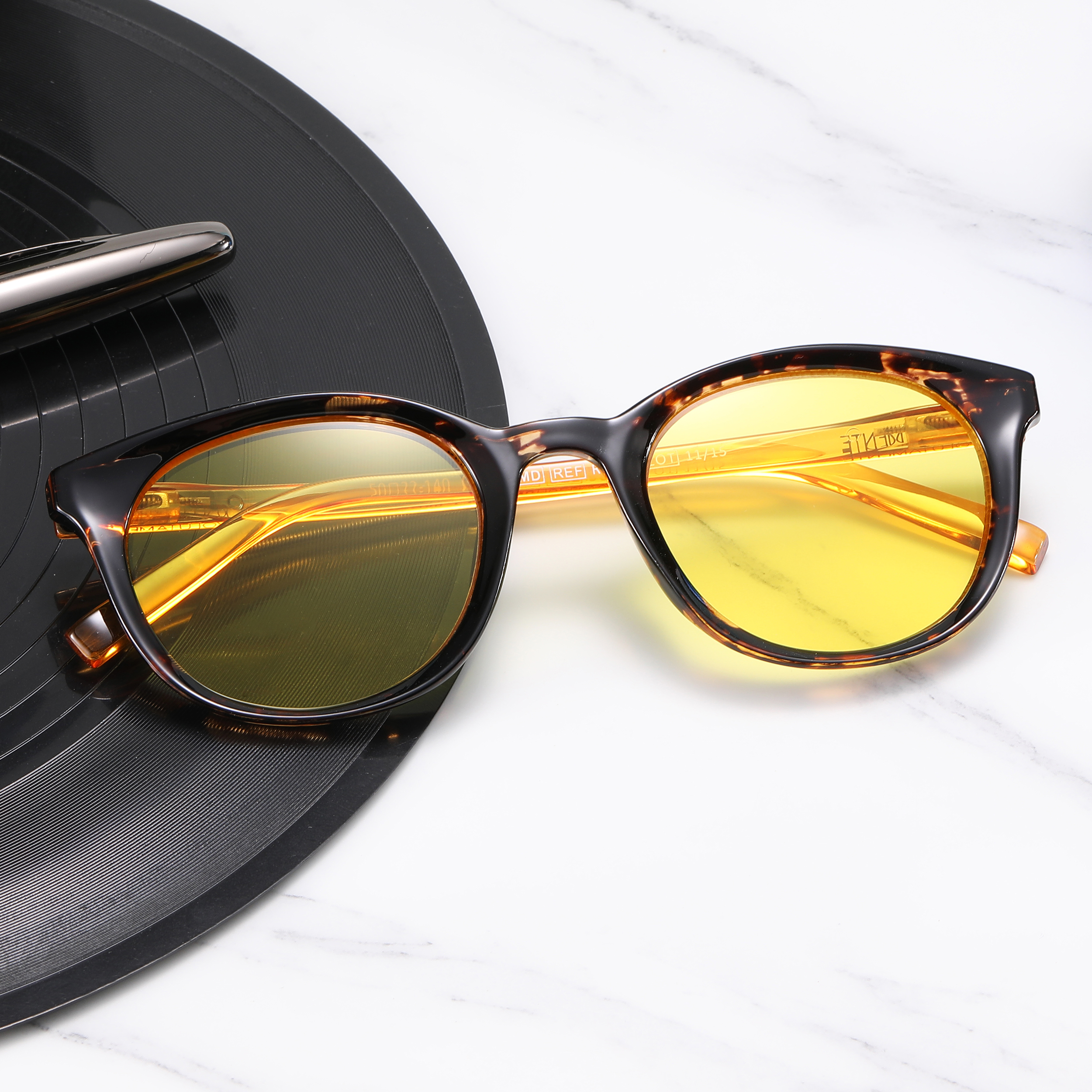 Close-up of tortoiseshell sunglasses with orange-yellow mirrored lenses resting on a marble surface beside a black circular object (lens cap).